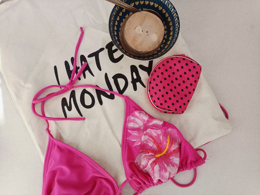 A flat lay arrangement featuring a bright pink bikini top with a floral print of a hibiscus flower in shades of pink and white, laid out on a white cloth with black text partially visible reading 'I H