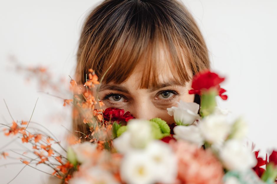 A woman with brown hair and striking blue eyes holds a bouquet of fresh flowers close to her face, partially obscuring her lower face and lips. The bouquet includes a variety of blooms such as white r