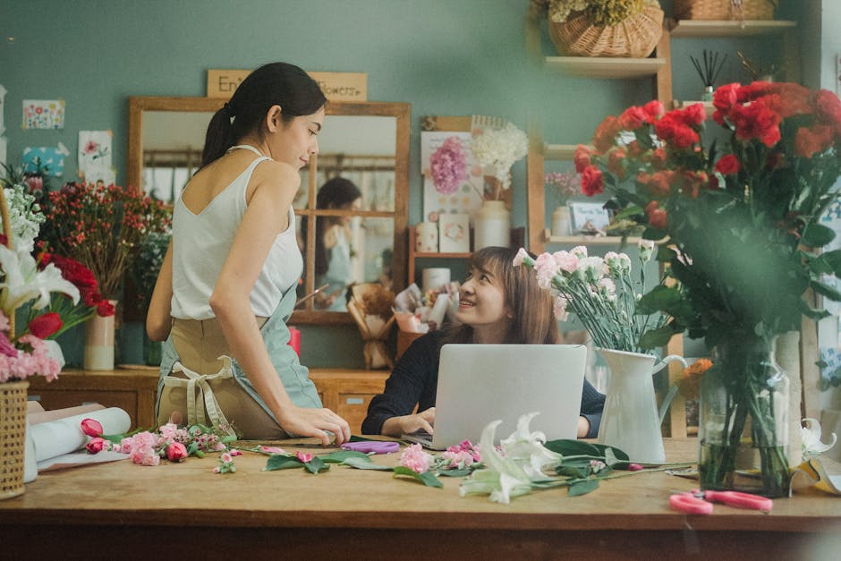 A female florist in a beige apron and white top is standing at a wooden table, engaged in a floral arrangement with pink and white flowers, including lilies and carnations, while smiling at a woman se