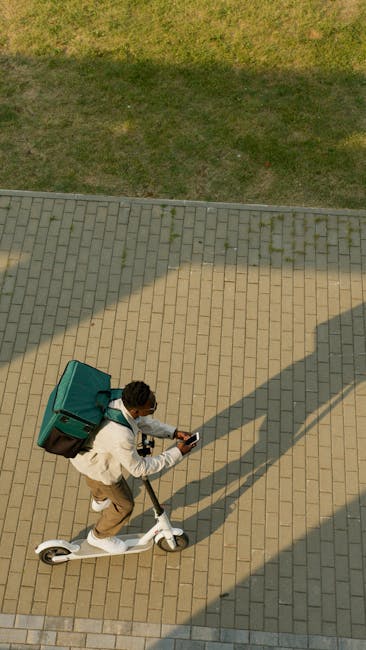 An aerial view of a young man riding a white electric scooter on a paved pathway outdoors during the daytime. He is wearing a white jacket and beige trousers, carrying a teal delivery box secured to h