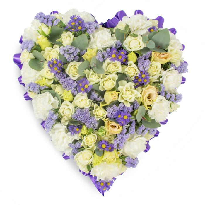 A lovingly assembled heart-shaped floral tribute presented on a plain white background, its outline precisely finished with a continuous band of darker purple satin ribbon that lends a refined edge to the whole design. Within that border the arrangement is densely composed of cream and soft yellow roses-many held in close, bud-like curls with some petals just beginning to open and revealing subtle green undertones-creating a plush, layered texture. Interleaved among those roses, full white carnations with ruffled edges build a soft, pillowy foundation. Delicate sprays of pale lavender statice drift across the surface, while groups of vivid purple, aster-like blooms with bright yellow centres inject small, joyful points of colour and eye-catching contrast. Scattered blue-green eucalyptus leaves, their round forms and muted sheen, break the floral density and introduce a cool, natural geometry that calms the composition. The shot is lit evenly, the soft light bringing out petal translucence and the satin ribbon's gentle sheen, suggesting a fresh, subtle fragrance of rose and eucalyptus. This arrangement reads as a serene, dignified offering-perfect for a funeral service in the Deptford area, a graveside tribute near Creek Road, or a chapel gathering by New Cross Gate-expressing consolation, enduring devotion, and measured sorrow. Its expert construction speaks to thoughtful timing and care, the kind of arrangement one would choose when seeking to honour and remember with quiet grace.