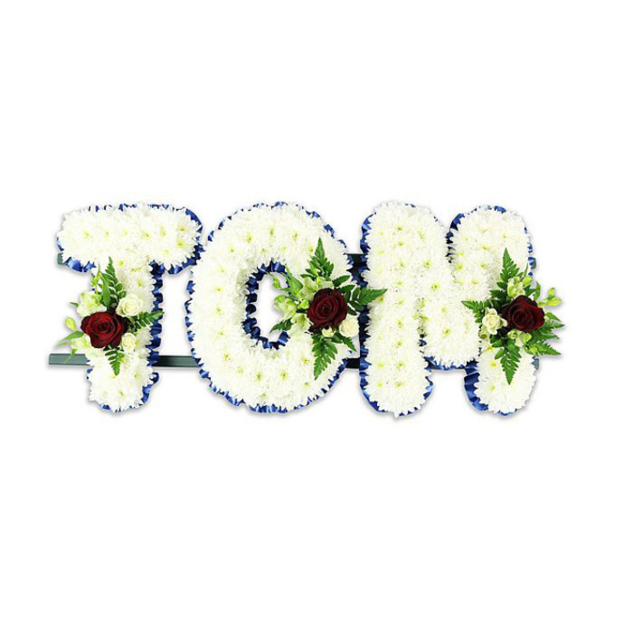 A personalised memorial display spells out 'TOM' in large, three-dimensional letters, each letter constructed from a dense mat of small, immaculate white chrysanthemums that produce a plush, cloud-like surface. The outer edge of every letter is neatly bordered with a royal blue ribbon, its rich tone providing a crisp profile against the white blooms and lending formal definition to the name. Within the body of each letter a deliberate floral spray has been positioned: a single, richly hued dark red rose dominates the cluster as an emblem of love and remembrance, while smaller creamy-white spray roses and fine fern fronds offer contrast, texture and a soft cascade of greenery. The overall balance is restrained and solemn, the red focal points offering emotional weight without disturbing the calm of the white chrysanthemum field. A narrow dark metal support is visible along the base, indicating the letters are intended to stand upright on an easel or near a headstone. Set against a plain white background in the picture, the arrangement's colours and detailed workmanship read clearly; in use, whether displayed inside a chapel on Deptford High Street or left near a village church, the composition would convey both dignity and intimacy. The scent is understated-clean chrysanthemum notes with the warmer, more resonant perfume of the red roses-while the texture invites a tactile imagination of soft petals and crisp ribbon. This is a classic funeral tribute, handcrafted to deliver a personalised message of farewell and solace for family and friends gathering to remember a loved one.