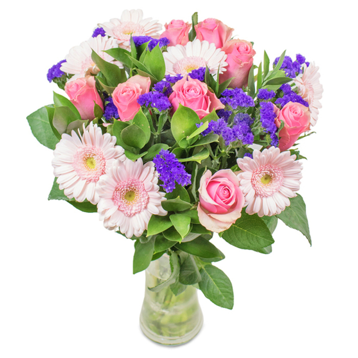 This vibrant bouquet, set against a luminous white backdrop, is a testament to the rich floral traditions and community spirit of Deptford. At its heart, 7-9 pink roses-some buds tightly coiled, others unfurling into elegant spirals-are interwoven with 5-7 airy pale pink gerbera daisies, their round faces and softly layered petals brightening the ensemble with a gentle, cheerful glow. Clusters of vivid purple statice punctuate the bouquet, adding both rich colour and intriguing texture reminiscent of the creative energy buzzing through Deptford's galleries and craft fairs. Lush green foliage forms an abundant base, reinforcing the freshness and beauty of each bloom, much like the thriving growth along the Thames Path. Arranged in a clear glass vase, the arrangement's natural aesthetic is enhanced by visible stems and glistening water, suggesting both impeccable freshness and the promise of same-day delivery to homes across Deptford. Ideal for birthdays, anniversaries, Mother's Day, or as a striking finishing touch in an office or front window, this bouquet of pink roses, gerberas, and purple filler radiates a spirit of celebration, affection, and Deptford's signature welcoming warmth.