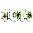 This photograph shows a personalised funeral flower arrangement spelling out TOM in bold, freestanding capital letters, created with care and attention to detail by a local Deptford florist. Each letter is completely covered with pristine white chrysanthemums, their small, rounded petals packed so closely together that they form a thick, cushion-like layer. The overall impression is one of softness and purity, as if the letters were carved from clouds. Encircling the outer edge of every letter is a royal blue ribbon, gently pleated to produce a subtle ruffle that catches the light. This ribbon border sharply defines the shape of the T, O, and M, adding a calm, dignified note of colour that balances the white chrysanthemums. On each letter, one thoughtfully composed spray of accent flowers stands out: velvety red roses, fully open and rich in tone, are accompanied by petite white or cream spray roses and slender, white blooms that resemble freesia in their graceful form. Surrounding these main flowers, sprigs of glossy foliage and airy fern fronds bring depth and a touch of natural wildness, softening the formal outline of the letters. The three floral letters are aligned horizontally on a slim, dark support bar that keeps them stable while remaining visually unobtrusive. Against the plain white background, every detail - from the delicate petals to the interplay of red, white, blue, and green - can be clearly appreciated. The tribute feels both strong and tender, a bespoke floral name display that would sit beautifully at a service in Deptford, perhaps near a coffin or memorial table, offering a final, loving farewell to Tom in a way that is both personal and visually striking.